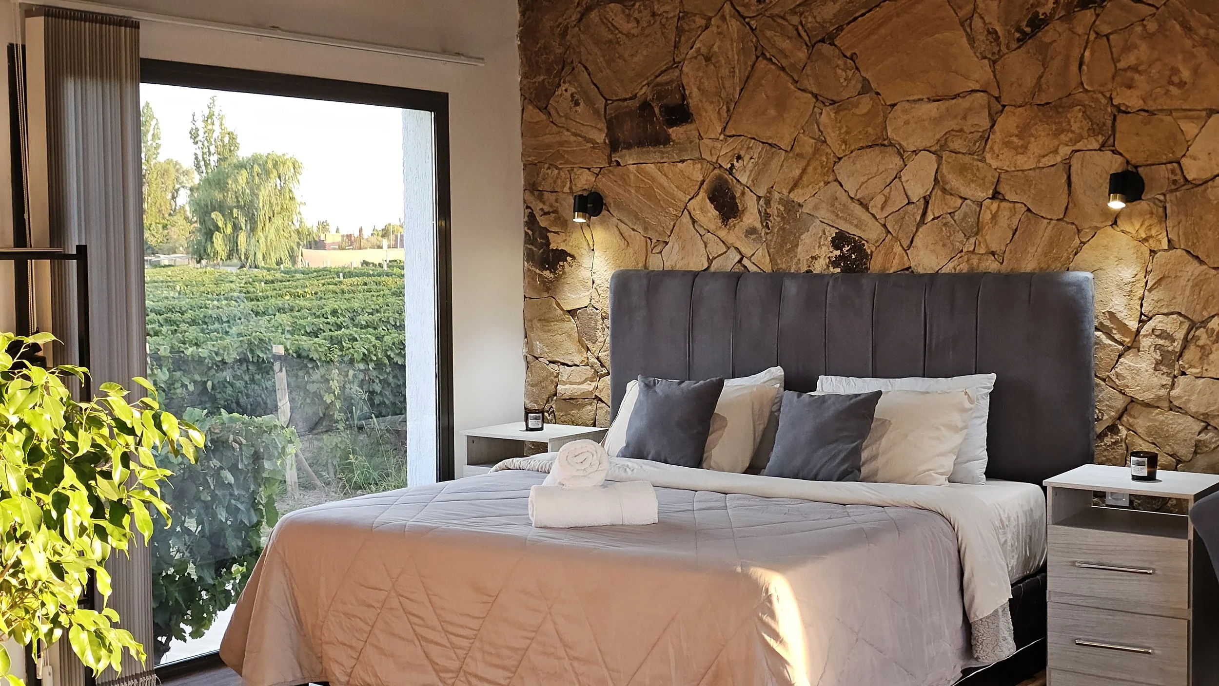 Dormitorio con una pared de piedra, cama con respaldo gris y almohadones blancos y grises. Dos mesas de luz con una vela cada una. Un ventanal grande deja ver los árboles de afuera y entra el sol. A la izquierda se asoma una planta.
