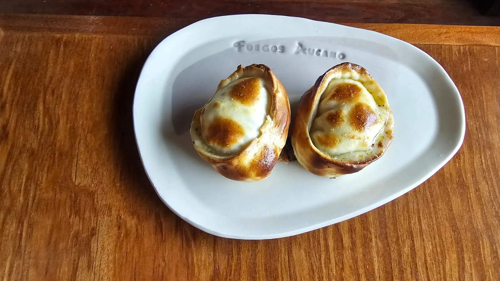 Dos huevos rellenos gratinados con queso, en un plato ovalado blanco con inscripción, sobre una mesa de madera.