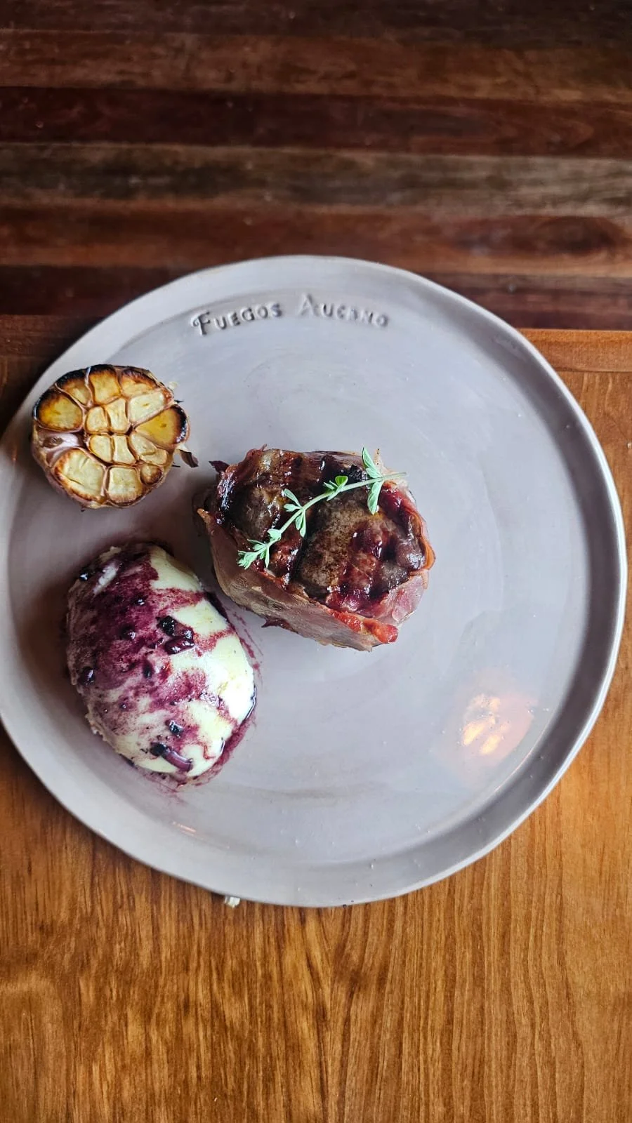 Un plato de cerámica con la inscripción "Fuegos Argentinos" con una porción de carne, una cabeza de ajo asada y una bocha de helado con salsa de frutos rojos, decorado con una ramita de hierbas.