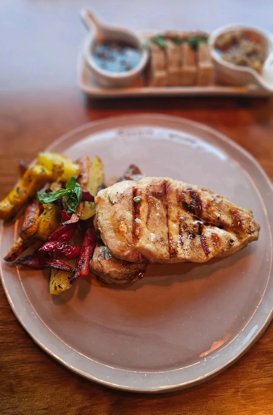 Pechuga de pollo grillada con verduras asadas en el plato y unos boles con salsas detrás.