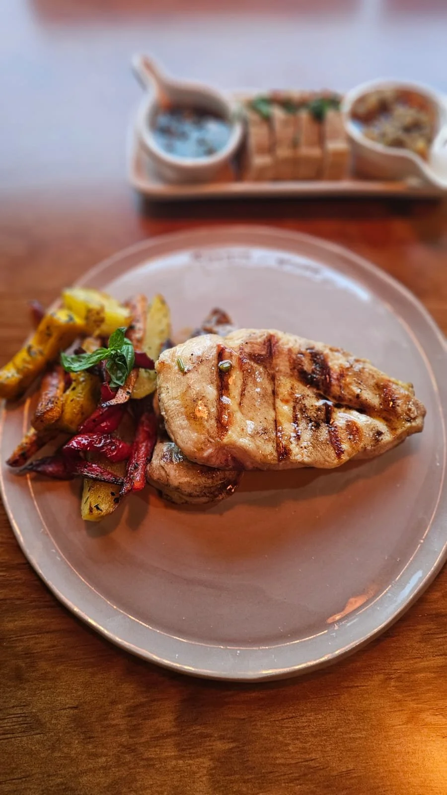 Pechuga de pollo grillada con verduras asadas en el plato y salsas para acompañar detrás.